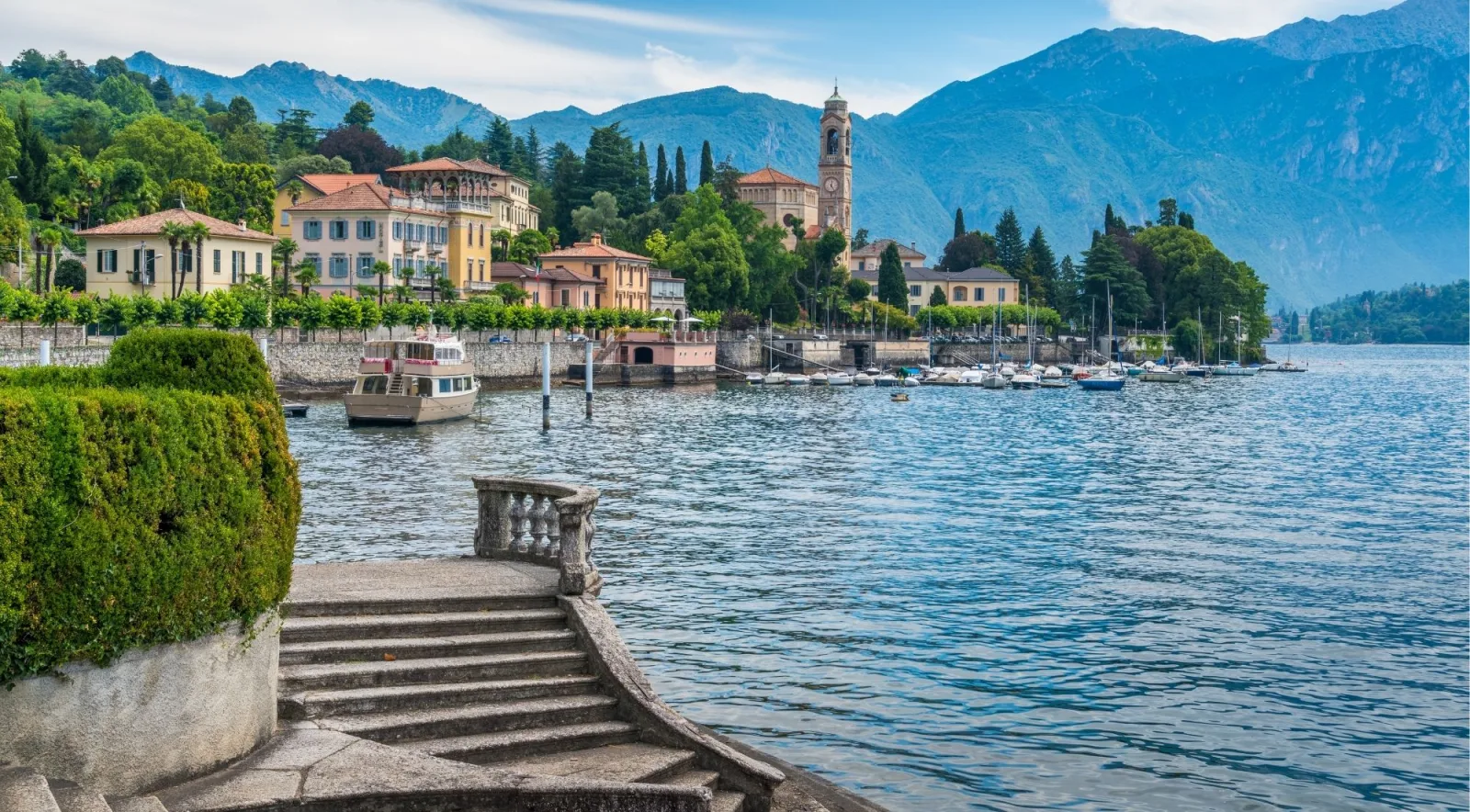 A lakeside village promenade on Lake Como with a bell tower, moored boats and stone steps down to the water
